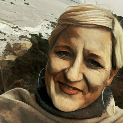 A photo of gs practitioner Sue Kuyper. A white person with short blond hair and wearing a grey sweater and silver hoop earrings is smiling into the camera. Behind her is the ocean and a rocky beach and a grey sky. The whole picture has a watercolor filter on top of it.