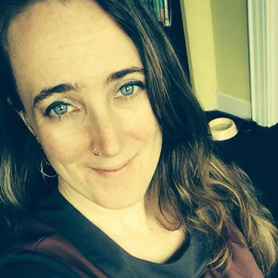 A photo of gs practitioner Becka Tilsen. A white woman with long wavy brown hair is looking and smiling into the camera. She is wearing a black sweater. Behind her is a white dog bowl and a wall painted yellow with white trim.