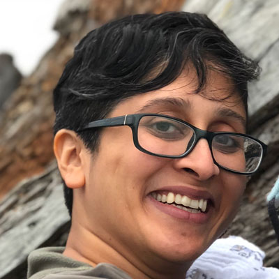 A photo of gs practitioner Bhavana Nancherla. A brown person with short salt and pepper grey and black hair is looing into the camera and smiling. They are wearing black-rimmed glasses and a green sweater and standing in front of grey and copper colored rock faces that are blurred in the background.