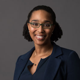A photo of gs practitioner Kira Banks. A black woman with black rimmed glasses and curly hair pulled back is smiling into the camera. She is wearing a blue suit and a necklace with a pendant on it and is standing infront of a dark grey background.