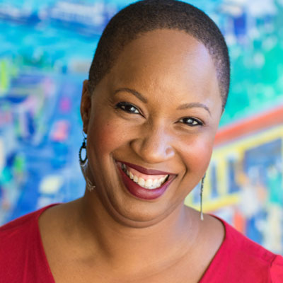 A photo of gs practitioner Sonya Brewer. A Black woman with short hair, dangly earrings, and wearing a red shirt with red lipstick is smiling into the camera. In the background is a multicolored wall, mostly shades of blue, some green, red, and yellow as well.