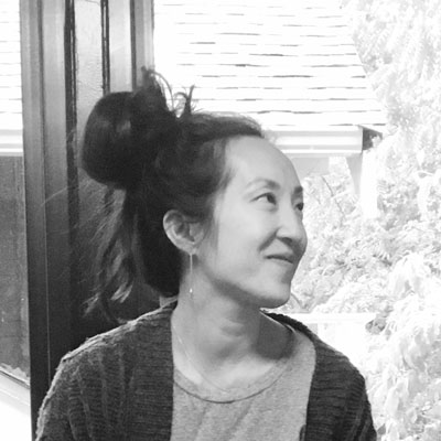 A photo of gs teacher Katelyn Song. A black and white photo of an Asian woman facing towards the right of the photo, wearing a dark cardigan over a light shirt with her long hair in a bun.
