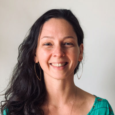 A photo of gs practitioner Becca Meredith. A white woman with long black hair, gold leaf-shaped hoop earrings, and a green shirt is smiling into the camera. Behind her is a white background.