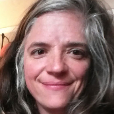 A photo of gs Practitioner Meredith Broome. A white woman with grey and black hair smiles into the camera, and behind her is a yellow and white wall.