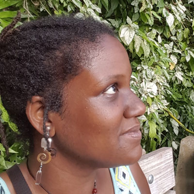 A photo of gs teacher and practitioner Lisa Thomas-Adeyemo. A black woman is looking up and to the right of the photo, with a smile on her face, her hair in dreadlocks, and wearing a dangly earring with a silver square, a copper spral, and a clear rectangle on it. Behind her is a bench and lush green bushes.