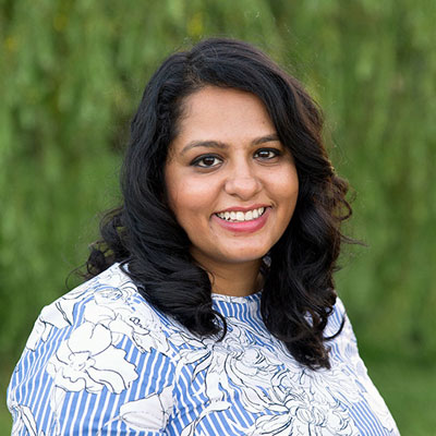 A photo of gs practitioner Piya Banerjee. A brown woman with long wavy black hair and a floral white and blue top is smiling into the camera, with a green background.