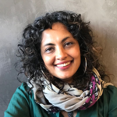 A photo of gs practioner Sumitra Rajukmar. A light-skinned brown woman with long curly black and brown hair, and wearing hoop earrings, a green top, and multi-colored scarf smiles into the camera.