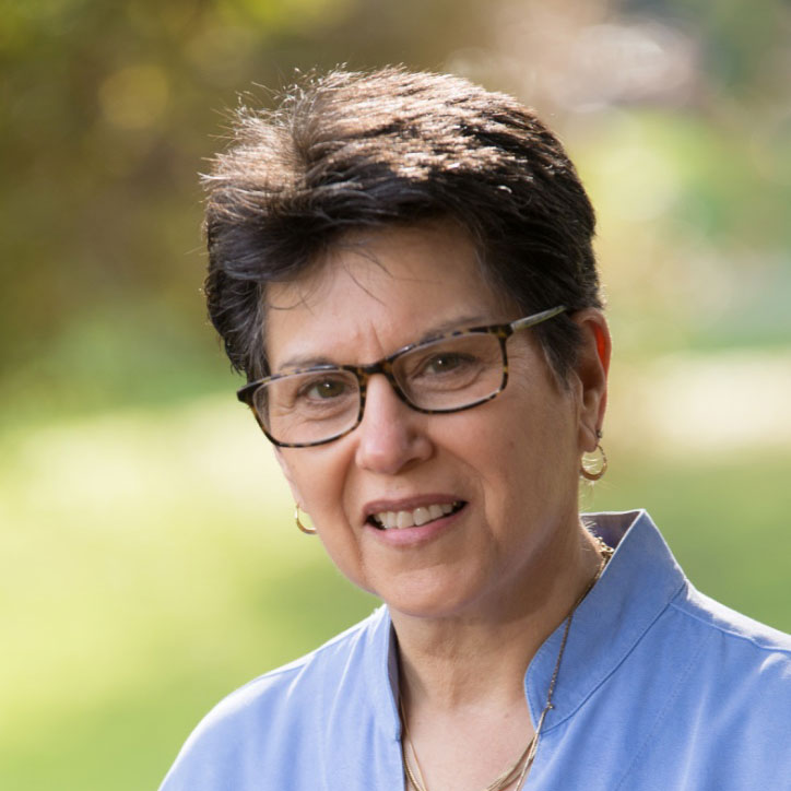 A photo of gs practitioner Denise Benson. A light-skinnned person with short hair and brown glasses is smiling into the camera. She is wearing a light blue shirt, and gold earrings and necklaces.