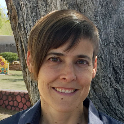 A photo of gs practitioner Jacks McNamara. A white person with asymmetrical brown hair is smiling into the camera. They are in front of a tree with grey bark and a wall and courtyard is behind them.