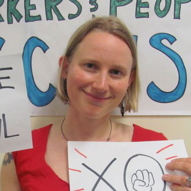 A photo of gs practitioner Clare Bayard, a white person with medium length blond hair smiling into the camera, wearing a red shirt, and holding up two hand drawn signs on paper.